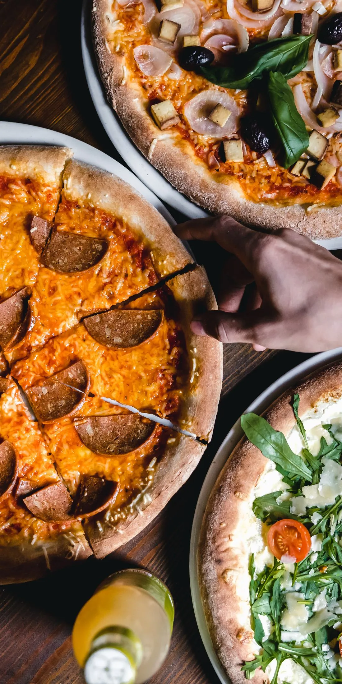 Drei Pizzen von oben auf einem Holztisch, zwei Hände nehmen sich ein Stück Salamipizza